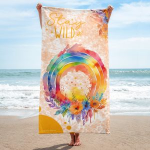 Chica con toalla multicolor Stay Wild en la playa. La toalla tiene un arcoíris multicolor con flores y plumas.