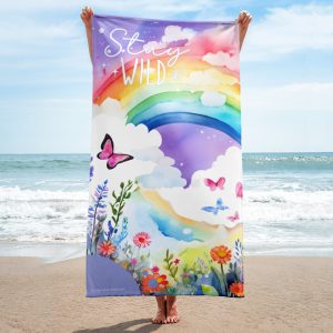 Chica con toalla multicolor con arcoíris, flores y mariposas Stay Wild en la playa